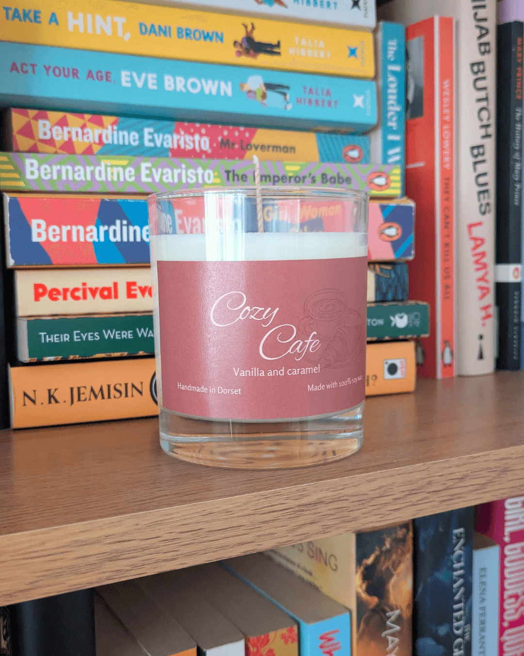 a soy candle with a brown label, reading 'Cozy Cafe'. The candle sits on a bookshelf, with a variety of books behind