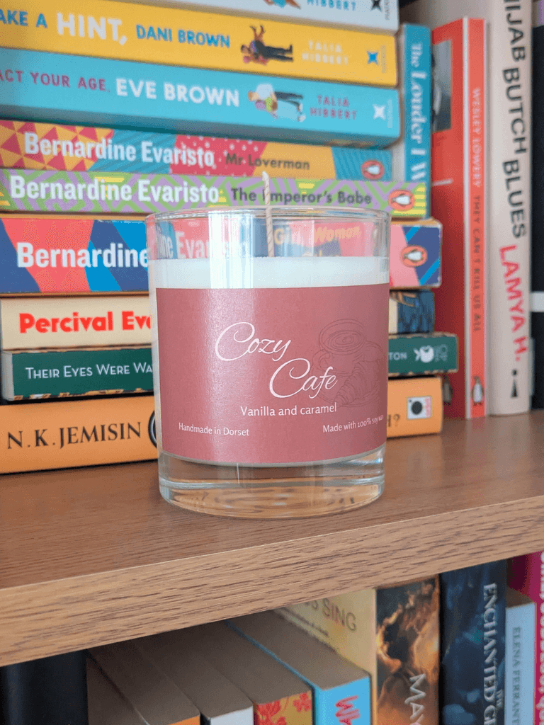 a soy candle with a brown label, reading 'Cozy Cafe'. The candle sits on a bookshelf, with a variety of books behind