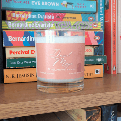 a soy candle with a pale orange label, reading 'Desert Mage'. The candle sits on a bookshelf in front of various books.