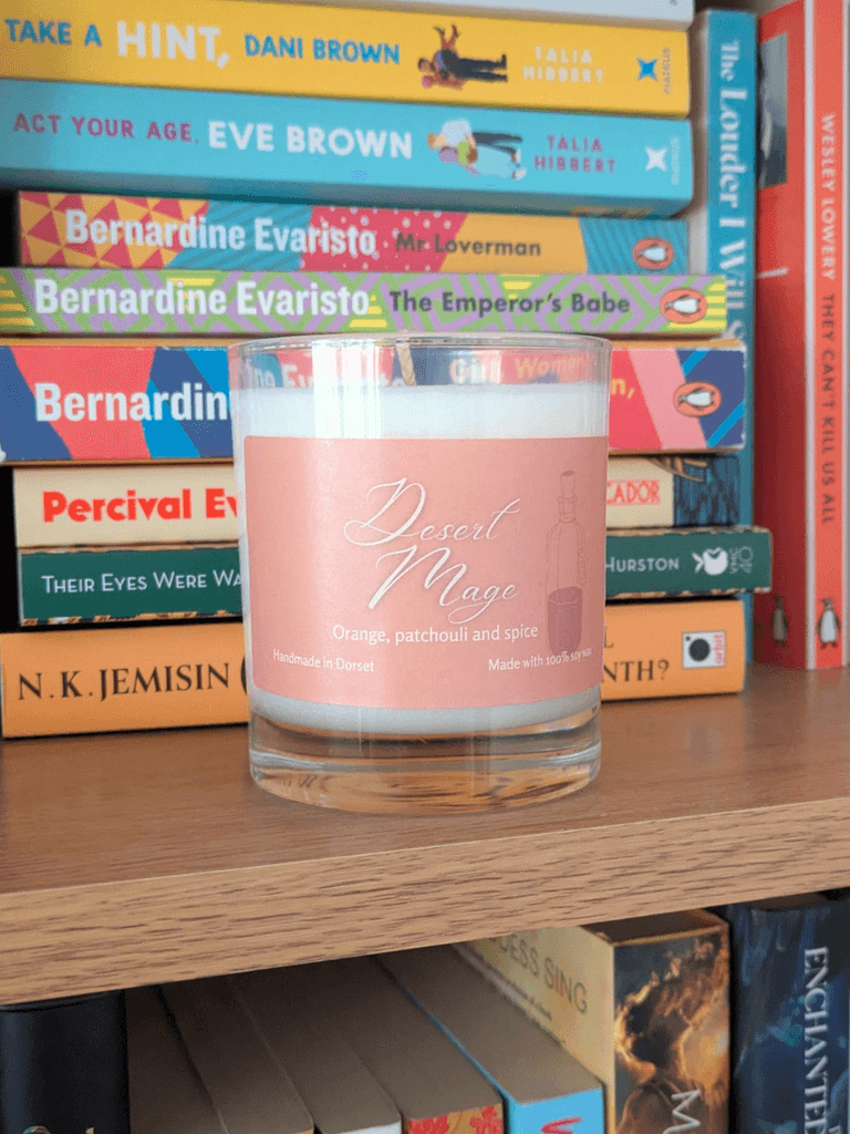 a soy candle with a pale orange label, reading 'Desert Mage'. The candle sits on a bookshelf in front of various books.