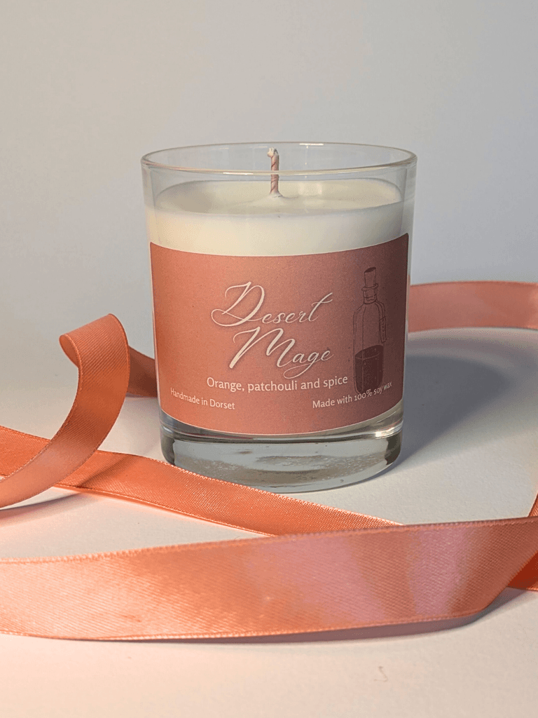a soy candle with a pale orange label, reading 'Desert Mage'. the table in front of the candle is decorated with a ribbon in the same orange shade.
