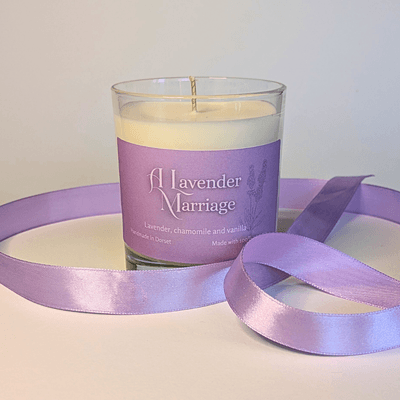 a soy candle with a lavender-purple label, reading 'A Lavender Marriage'. the table in front of the candle is decorated with a ribbon in the same lavender shade.
