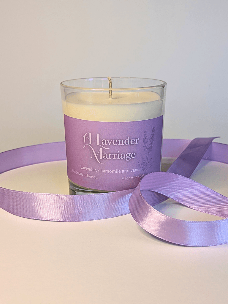 a soy candle with a lavender-purple label, reading 'A Lavender Marriage'. the table in front of the candle is decorated with a ribbon in the same lavender shade.