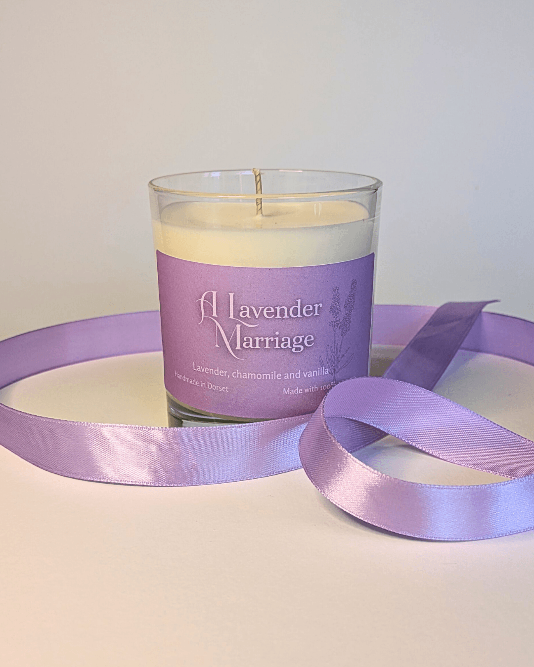a soy candle with a lavender-purple label, reading 'A Lavender Marriage'. the table in front of the candle is decorated with a ribbon in the same lavender shade.