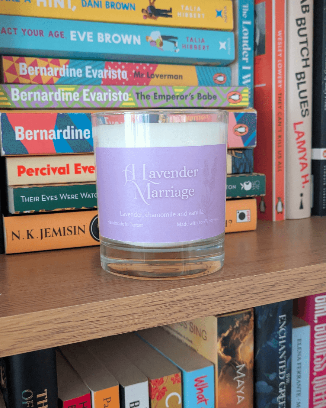 a soy candle with a lavender-purple label, reading 'A Lavender Marriage'. The candle sits on a bookshelf.= in front of various books.