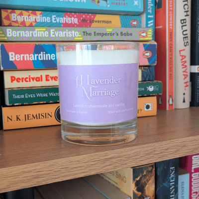 a soy candle with a lavender-purple label, reading 'A Lavender Marriage'. The candle sits on a bookshelf.= in front of various books.
