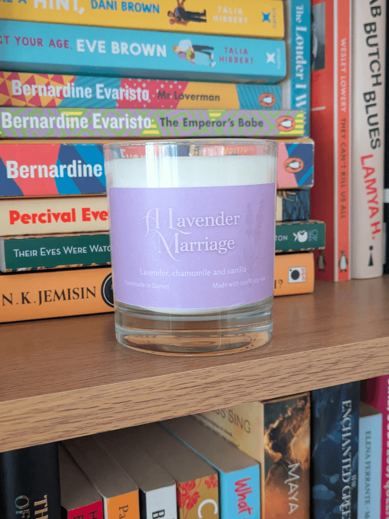a soy candle with a lavender-purple label, reading 'A Lavender Marriage'. The candle sits on a bookshelf.= in front of various books.