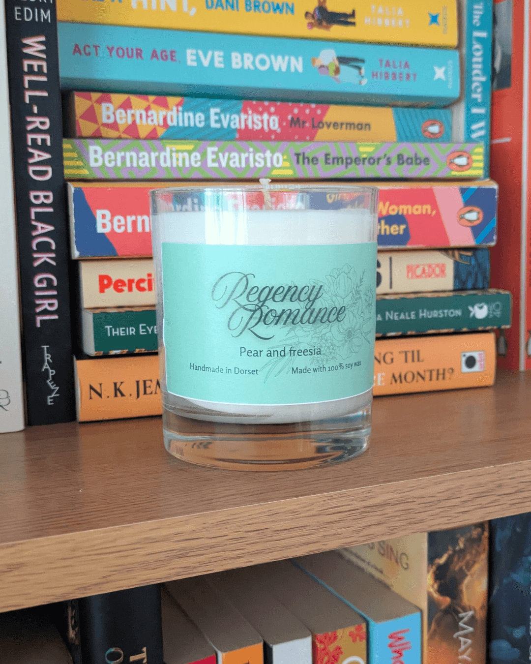 a soy candle with a mint-green label, reading 'Regency Romance'. The candle sits on a bookshelf in front of various books.