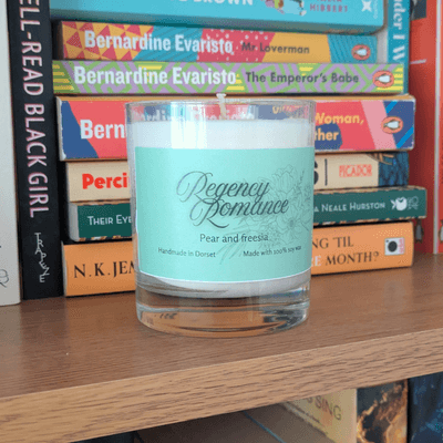 a soy candle with a mint-green label, reading 'Regency Romance'. The candle sits on a bookshelf in front of various books.