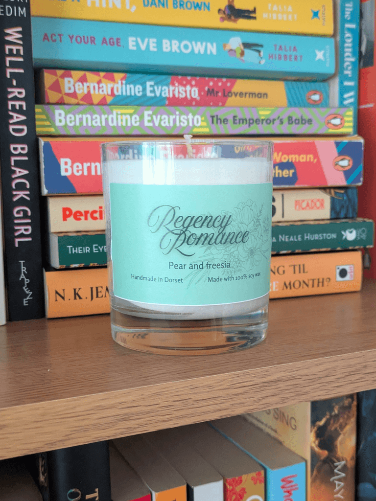 a soy candle with a mint-green label, reading 'Regency Romance'. The candle sits on a bookshelf in front of various books.