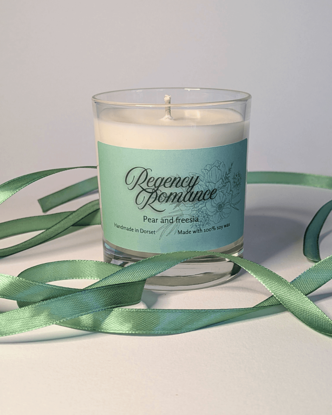 a soy candle with a mint-green label, reading 'Regency Romance'. the table in front of the candle is decorated with a ribbon in the same mint shade.